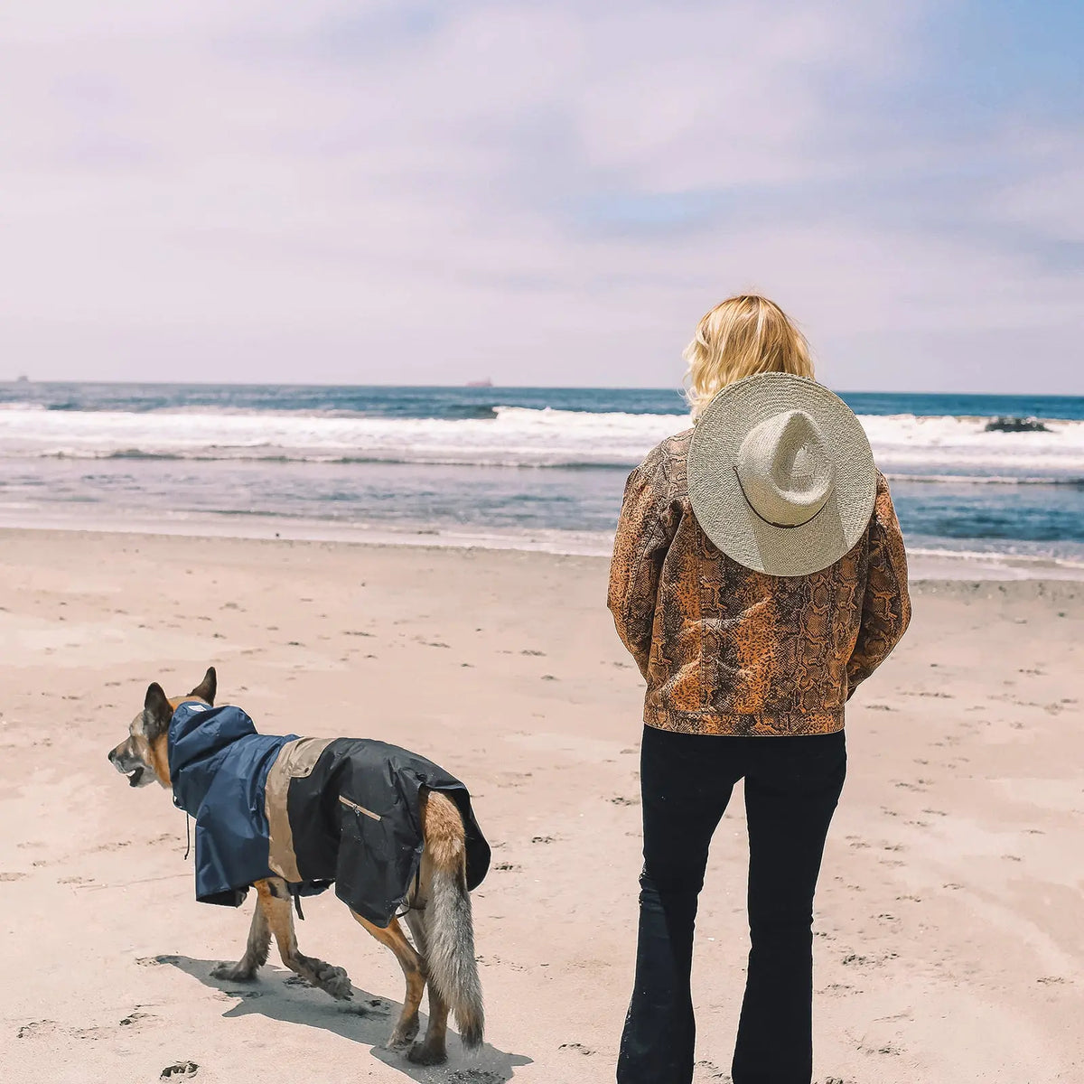 Chien portant le poncho imperméable Baja Coastline, debout sur la plage avec son maître et contemplant l’océan, prêt pour toutes les aventures côtières.