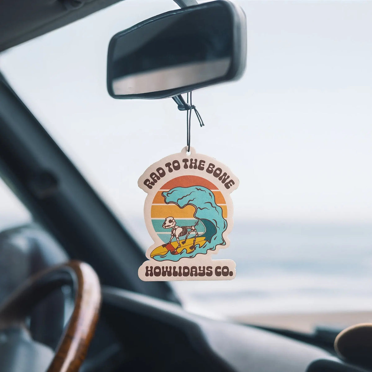Le Rad to the Bone Air Freshener suspendu au rétroviseur intérieur d’un véhicule, avec la plage en arrière-plan.